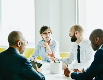 4 persons in an office, one of them is holding a tablet, papers are on the desk 4 persons in an office, one of them is holding a tablet, papers are on the desk